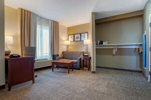 A hotel room with a sofa, a small table, a desk with a lamp, a chair, and a window with curtains. There are framed pictures on the wall and a shelf with items.