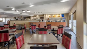 Um restaurante amplo, com pé direito alto, diversas mesas e cabines para refeições, área de cozinha e grandes janelas que proporcionam iluminação natural.