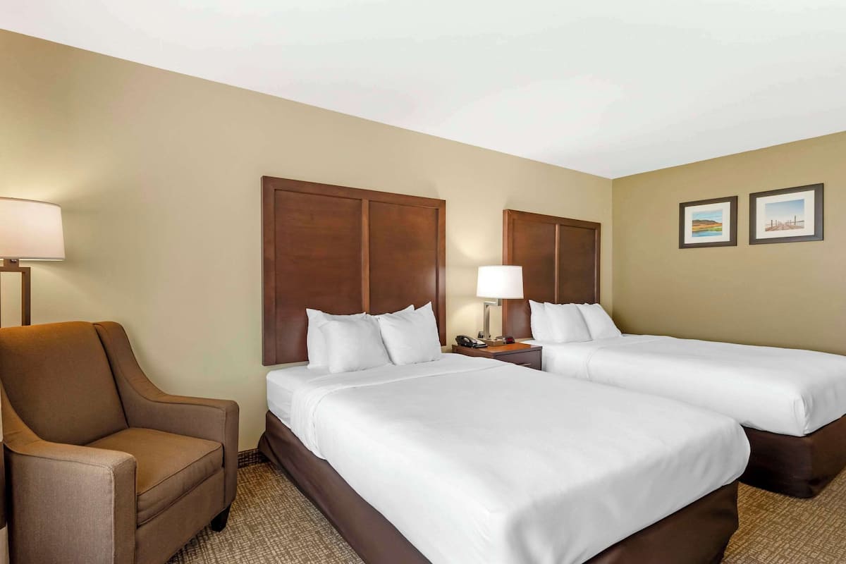 A hotel room with two beds, a chair, a nightstand with a lamp, and two framed pictures on the wall.