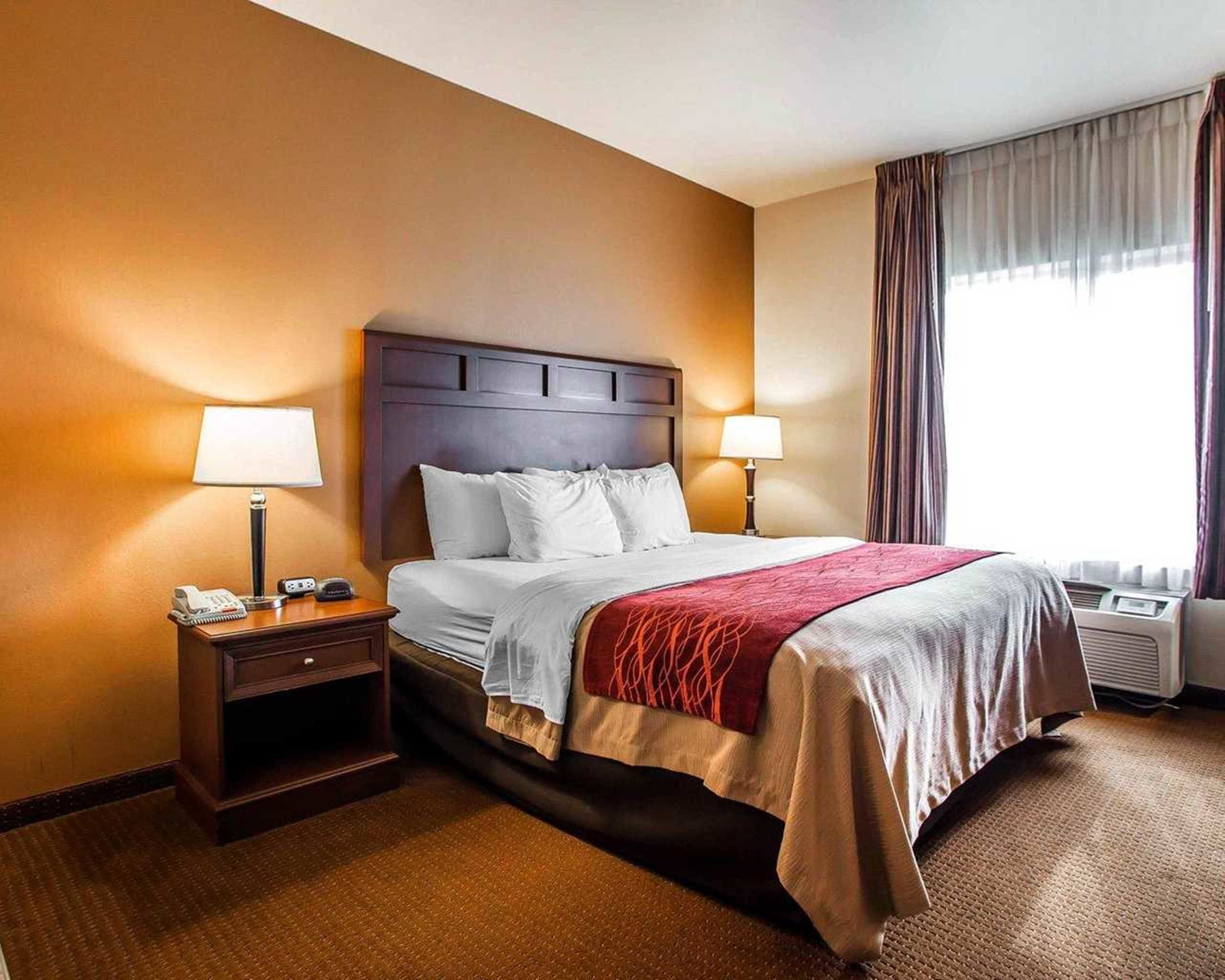 A hotel room with a large bed, two bedside tables, a nightstand, a window with curtains, and an air conditioning unit.