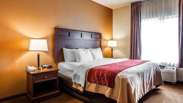 A hotel room with a large bed, two bedside tables, a nightstand, a window with curtains, and an air conditioning unit.