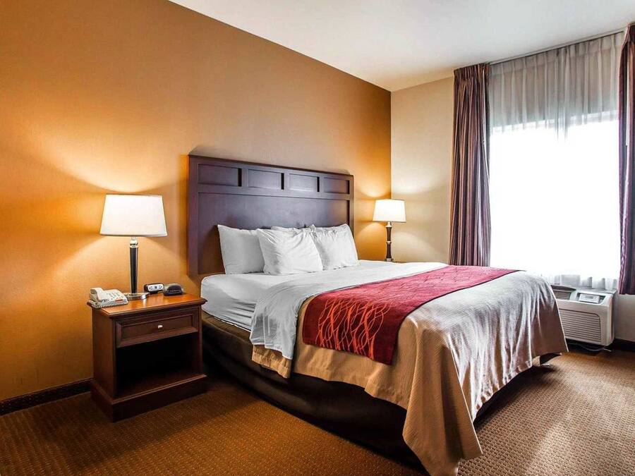 A hotel room with a large bed, two bedside tables, a nightstand, a window with curtains, and an air conditioning unit.