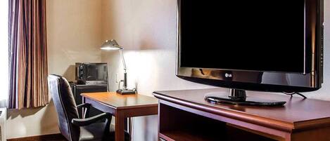 A hotel room with a flat-screen TV, a desk with a lamp, a chair, and a small desk lamp.