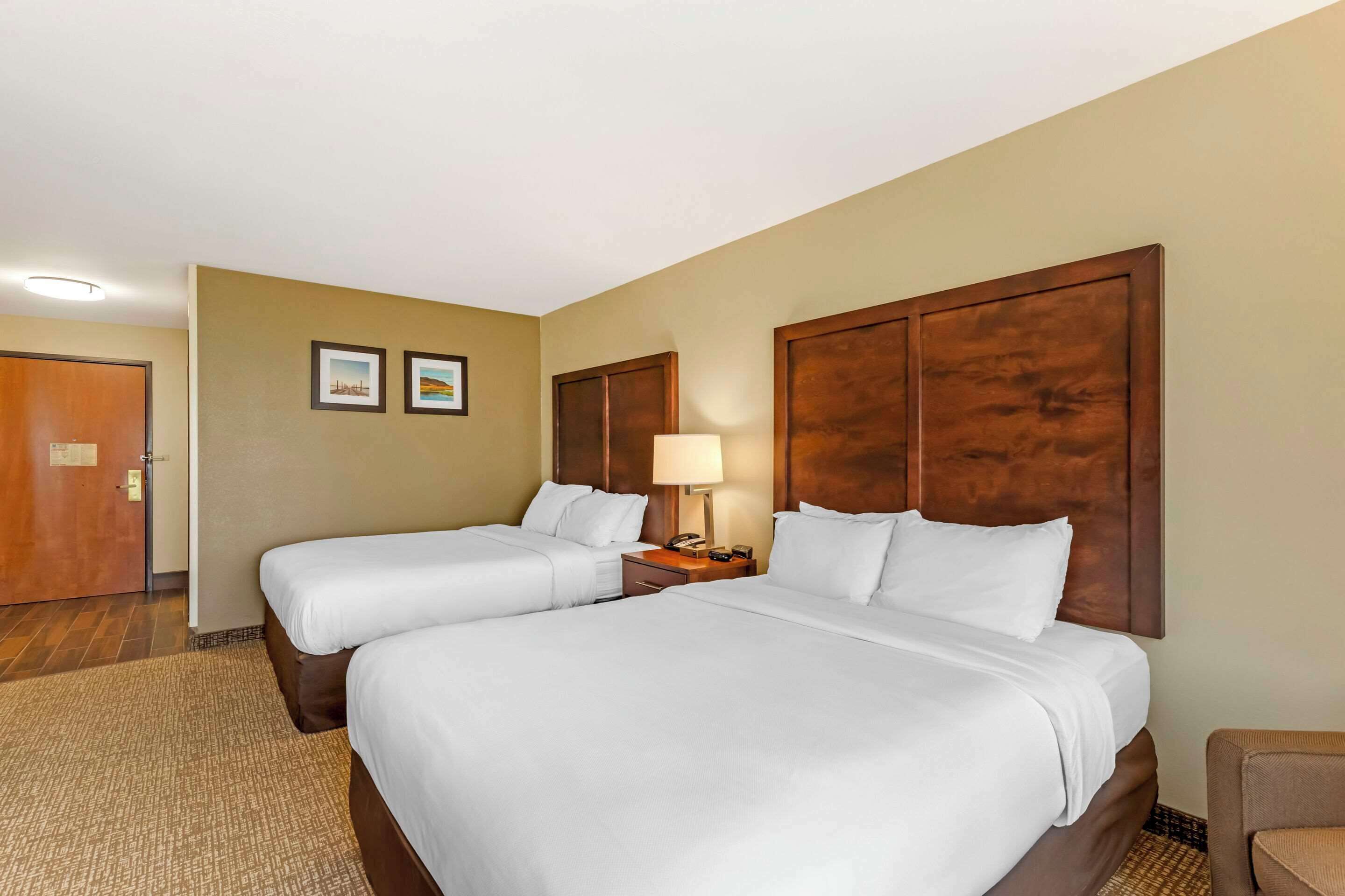 A hotel room with two beds, a nightstand with a lamp, a wooden headboard, and a door.