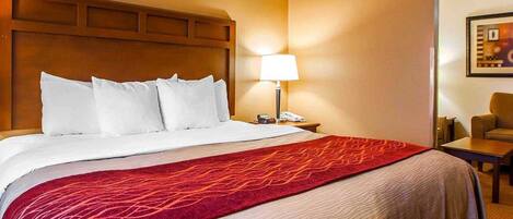 A hotel room with a large bed, a wooden headboard, a bedside table with a lamp, a chair, and a framed picture on the wall.