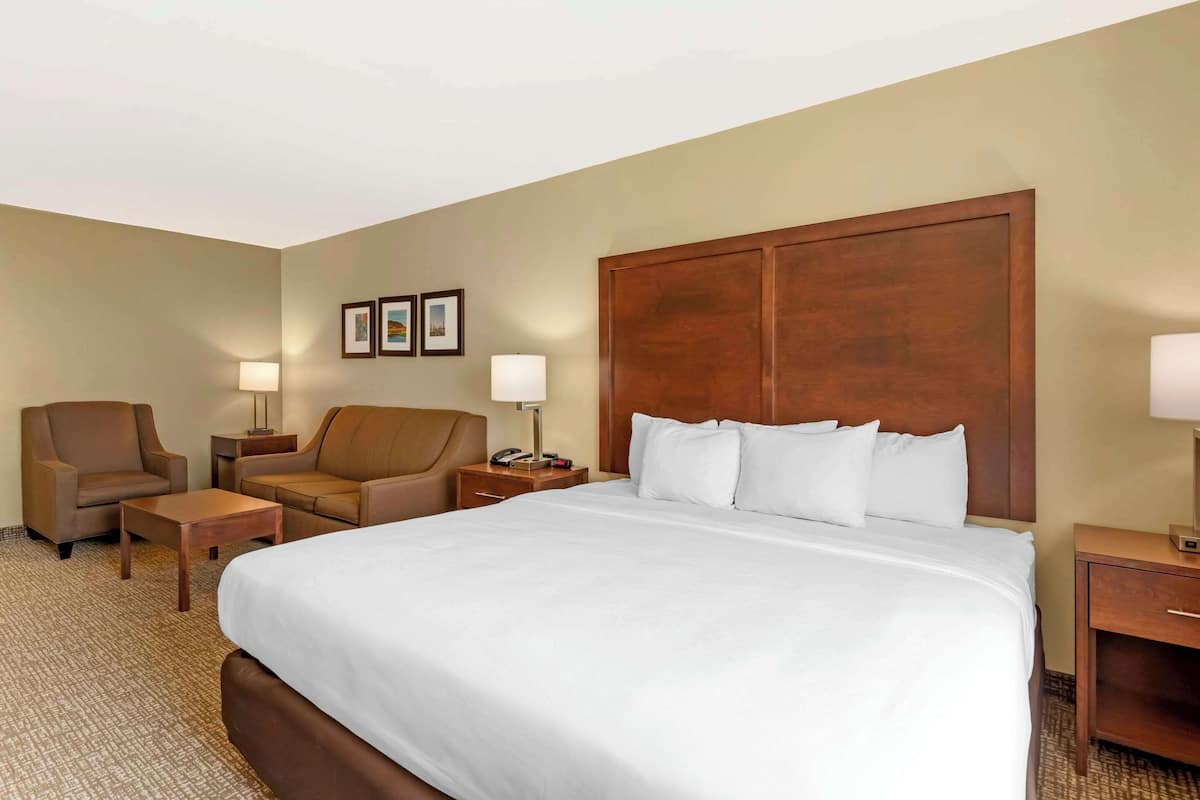 A hotel room with a large bed, two armchairs, a small table, a nightstand with a lamp, and a wooden headboard.