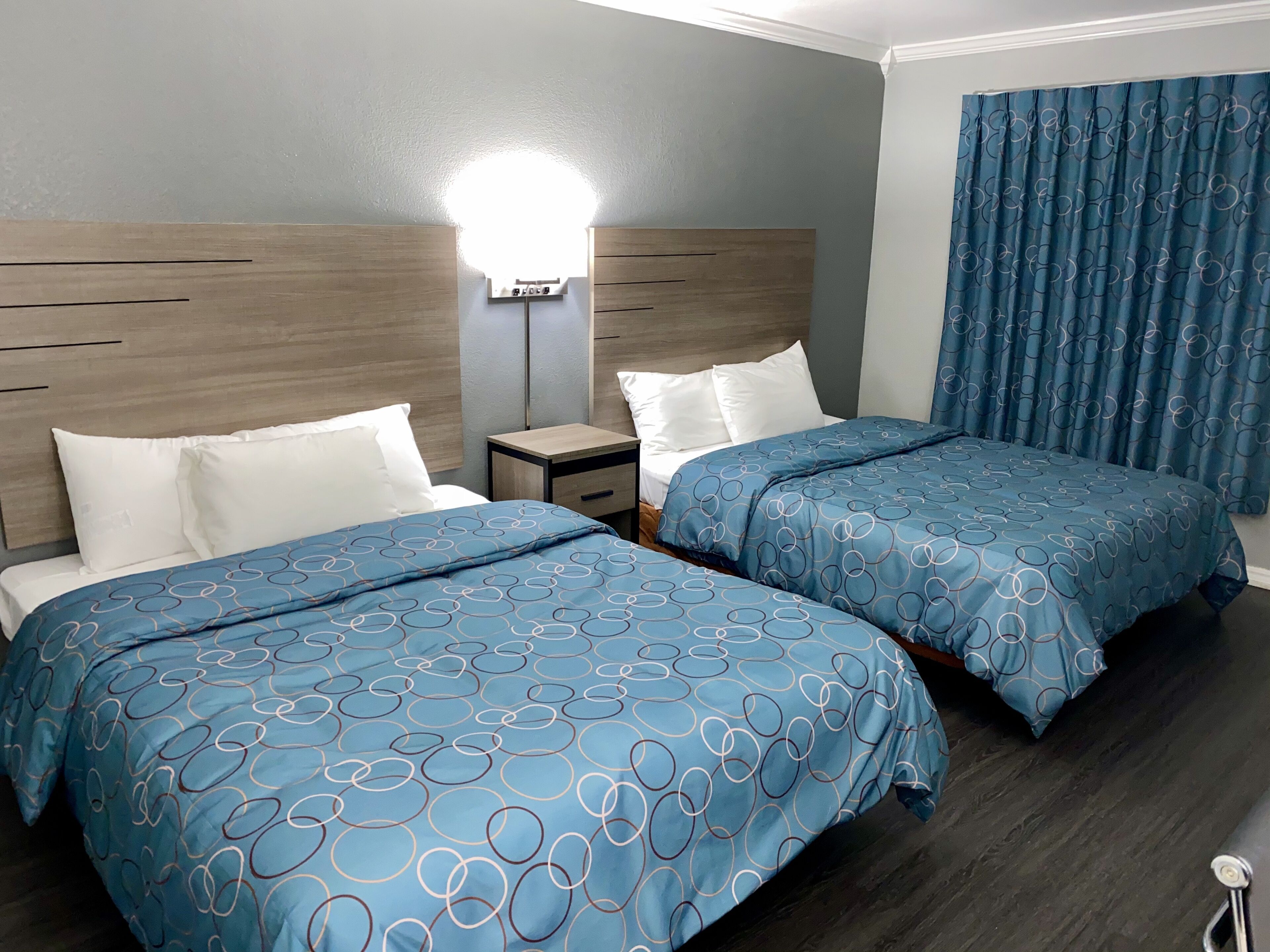 Una camera d'albergo con due letti, ognuno con lenzuola a motivi circolari blu, una testata in legno, un comodino con una lampada e una finestra con tende blu.