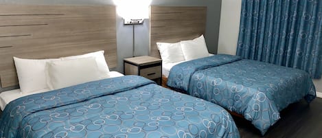 A hotel room with two beds, each with blue circular-patterned bedding, a wooden headboard, a bedside table with a lamp, and a window with blue curtains.