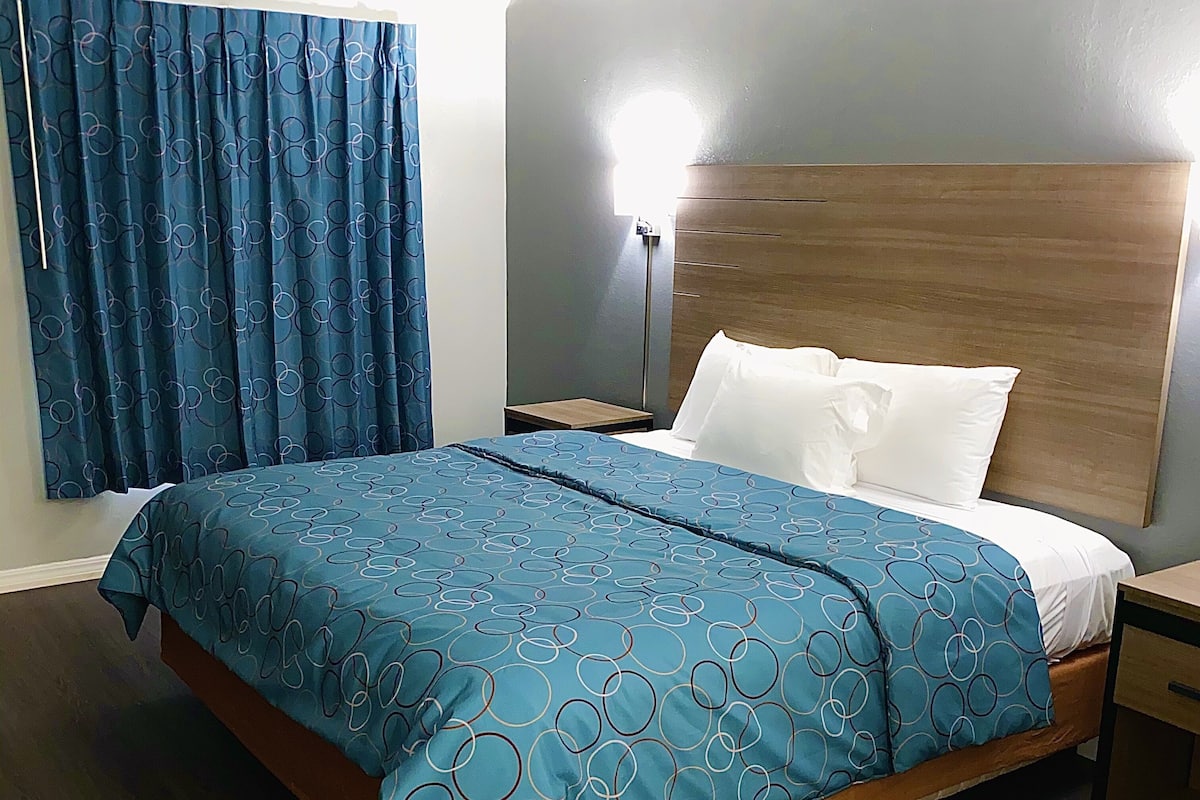 A hotel room with a bed, a nightstand, a lamp, and a window with curtains.