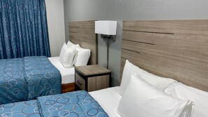 A hotel room with two beds, each with blue bedding and white pillows. There is a wooden headboard, a bedside table with a lamp, and a curtain with a circular pattern. - Budget Host Inn (Vernon)