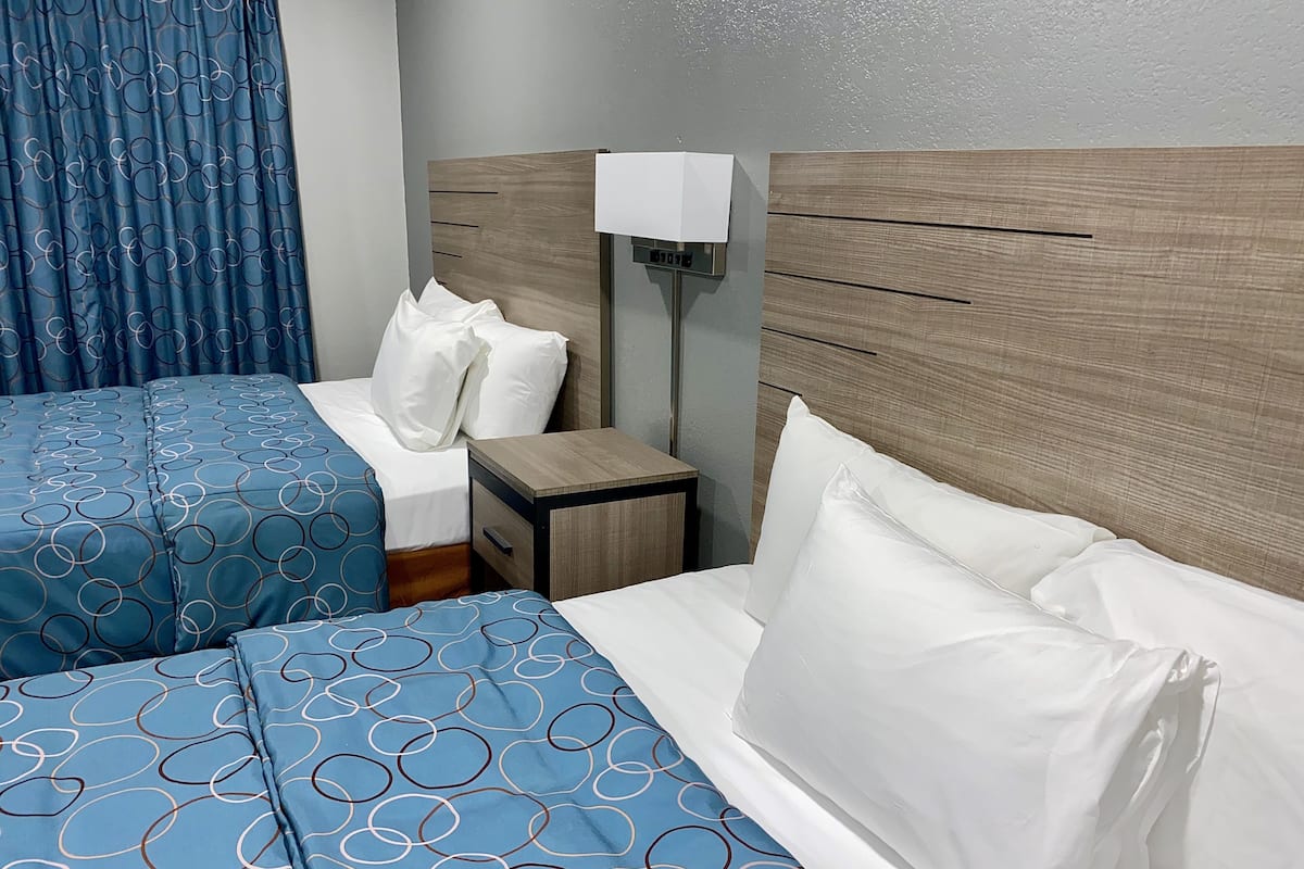 A hotel room with two beds, each with blue bedding and white pillows. There is a wooden headboard, a bedside table with a lamp, and a curtain with a circular pattern.