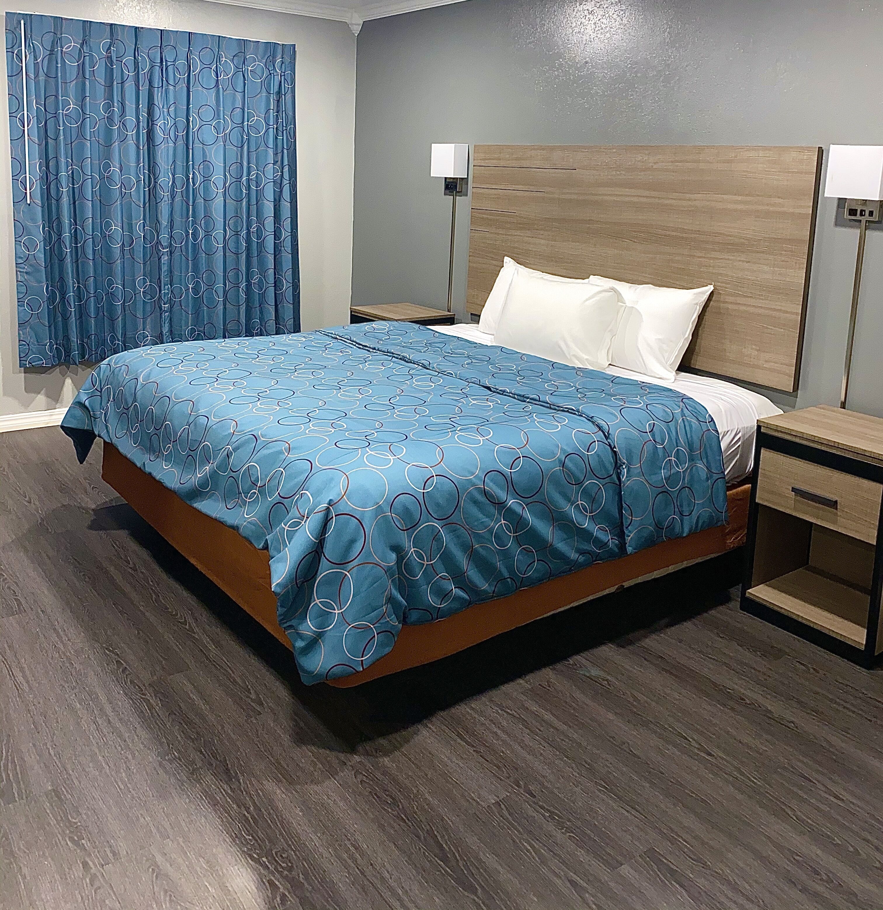 A hotel room with a bed, wooden headboard, nightstands, and a blue curtain.