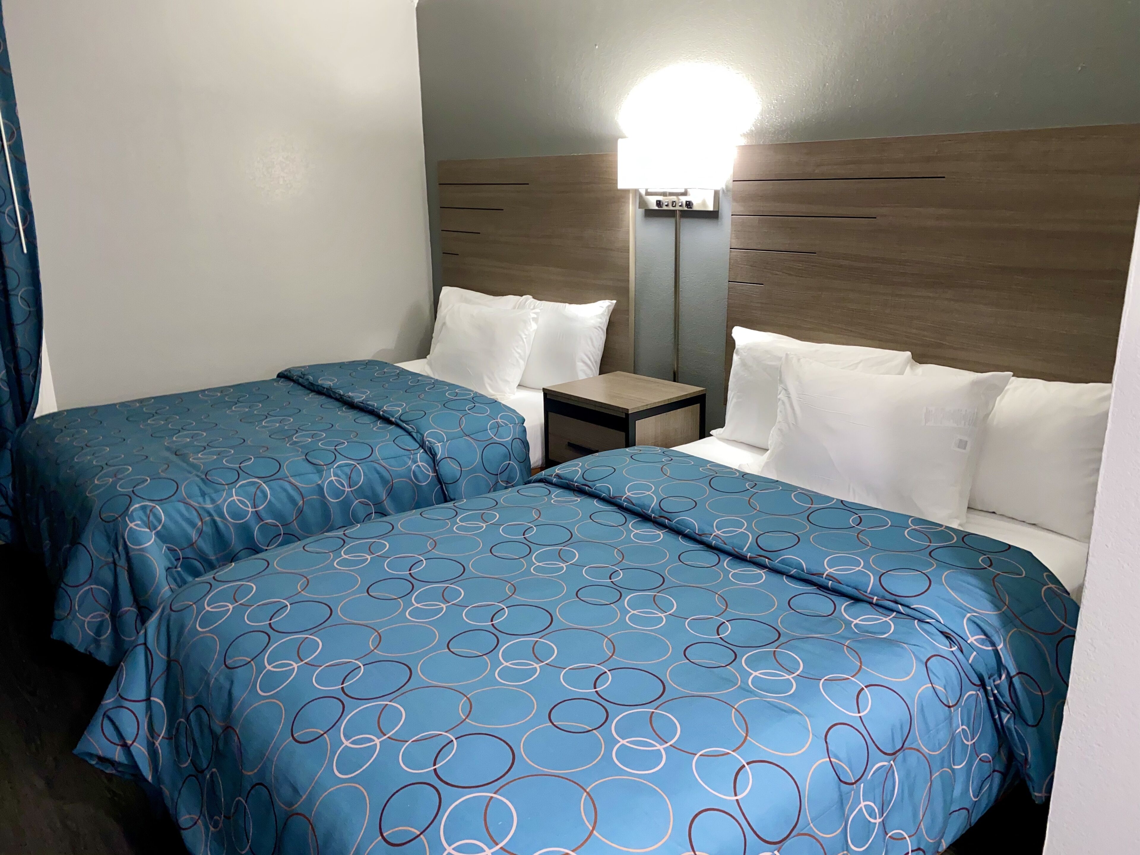 A hotel room with two beds, each with blue bedding featuring circular patterns. There is a bedside table with a lamp between the beds.