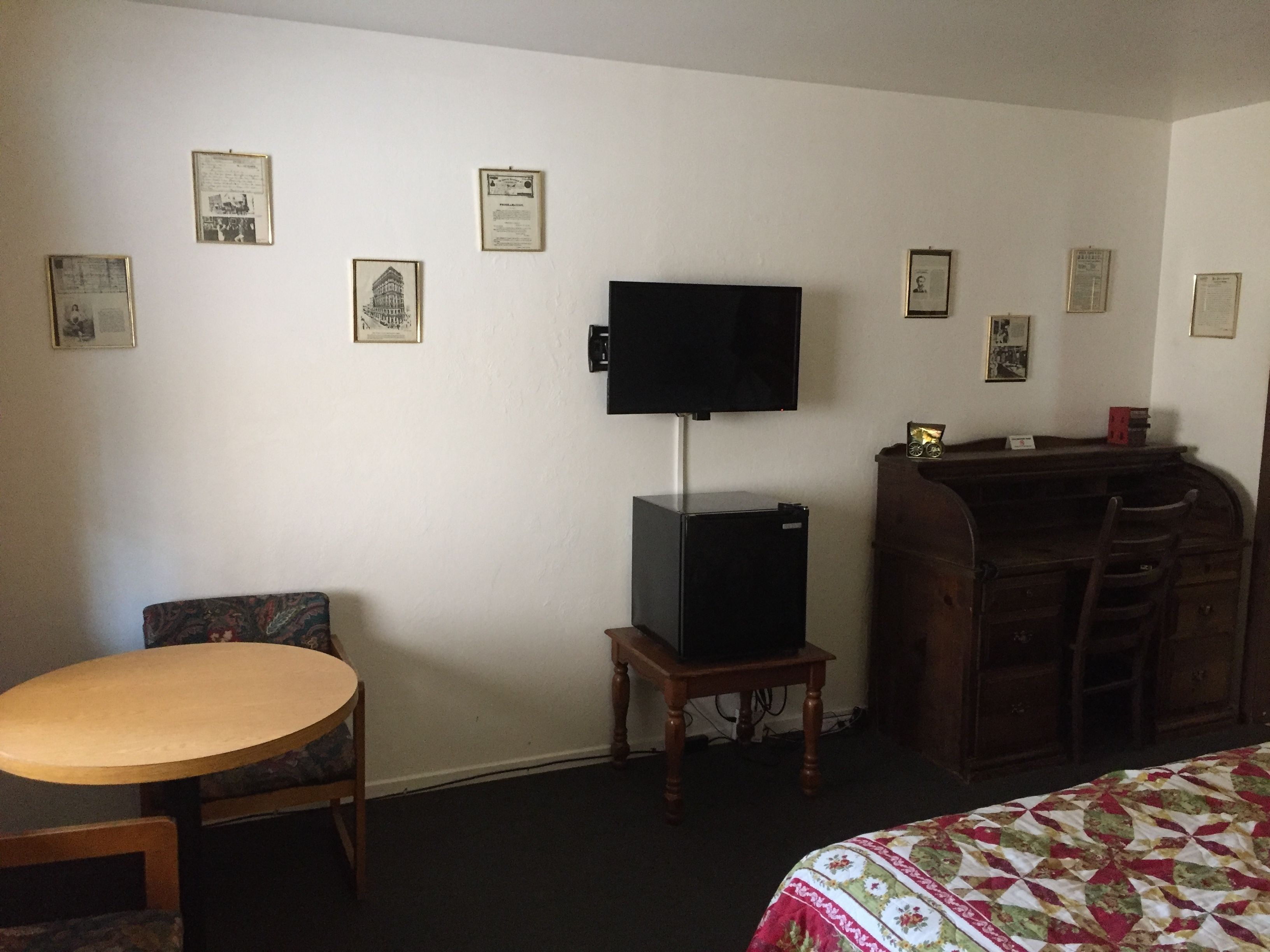 Une chambre d’hôtel avec un lit, un bureau, une chaise, un téléviseur posé sur un meuble et plusieurs tableaux encadrés accrochés au mur.