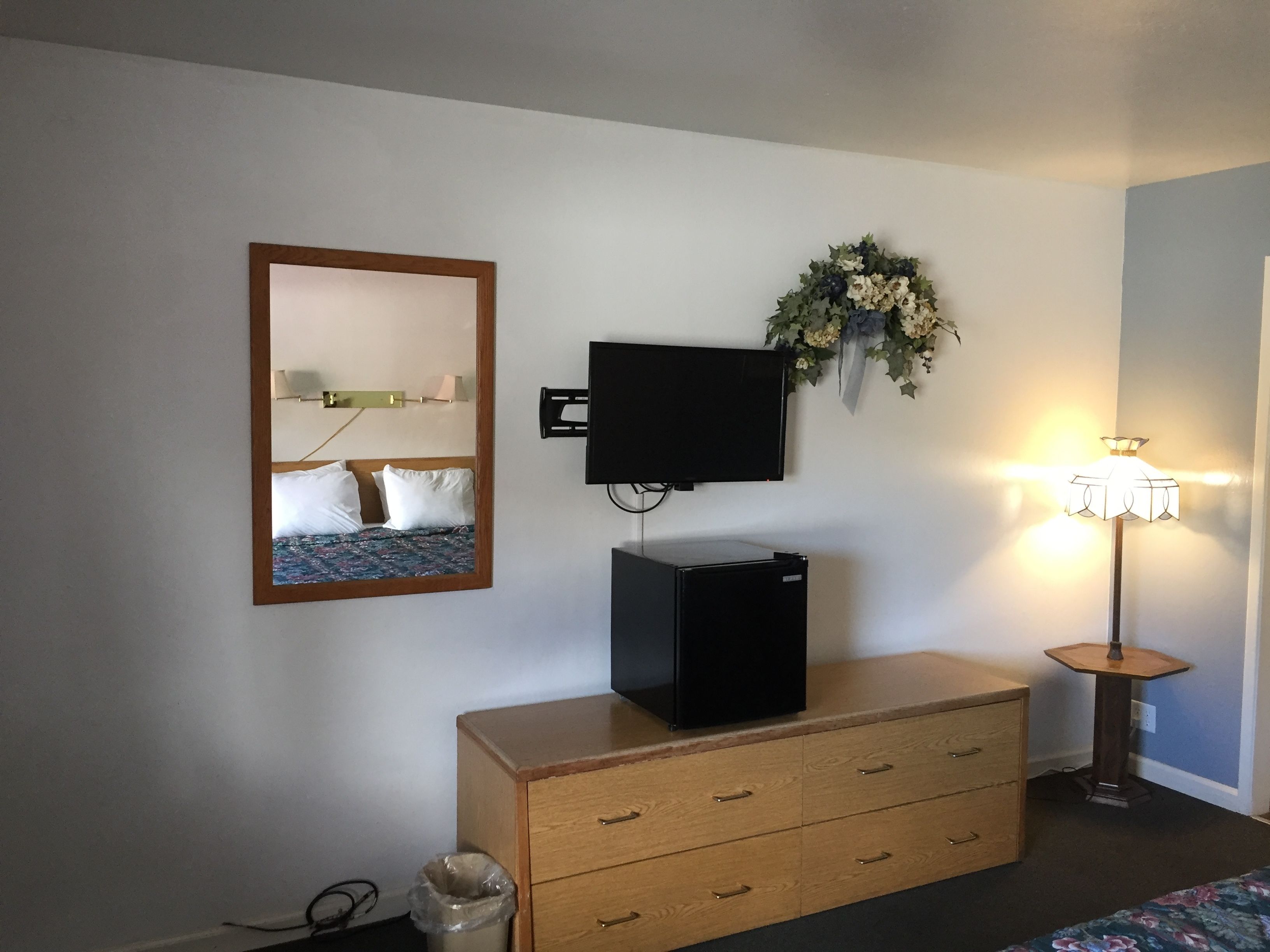 A hotel room with a bed, a dresser, a television, a wall-mounted mirror, a decorative wreath, and a floor lamp.