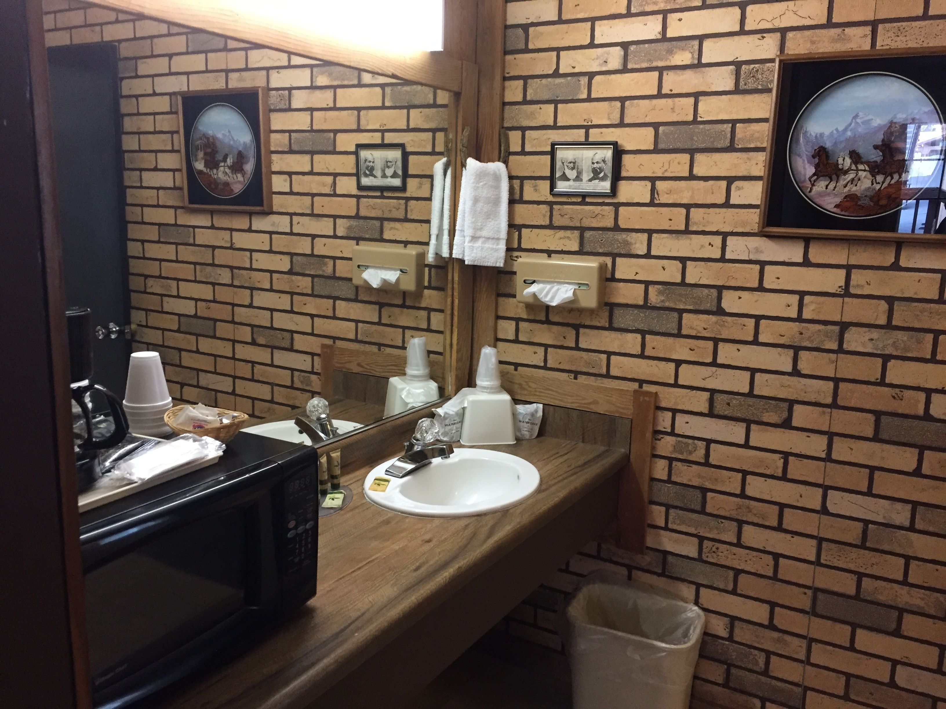A bathroom with a wooden vanity, a sink, a mirror, and a trash bin. There are framed pictures on the wall and a towel dispenser.