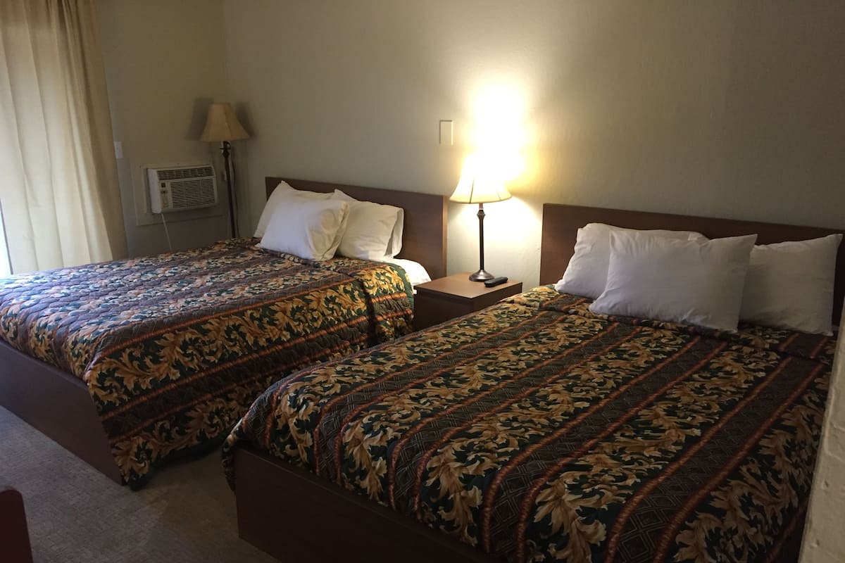 A hotel room with two beds, each with a patterned bedspread, a nightstand with a lamp, and a wall-mounted air conditioning unit.