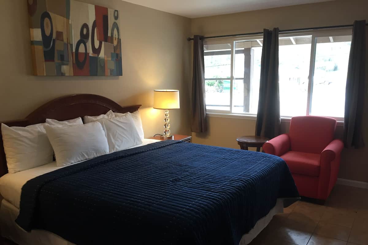A hotel room with a large bed, a red armchair, a small table, a lamp, and a window with curtains.