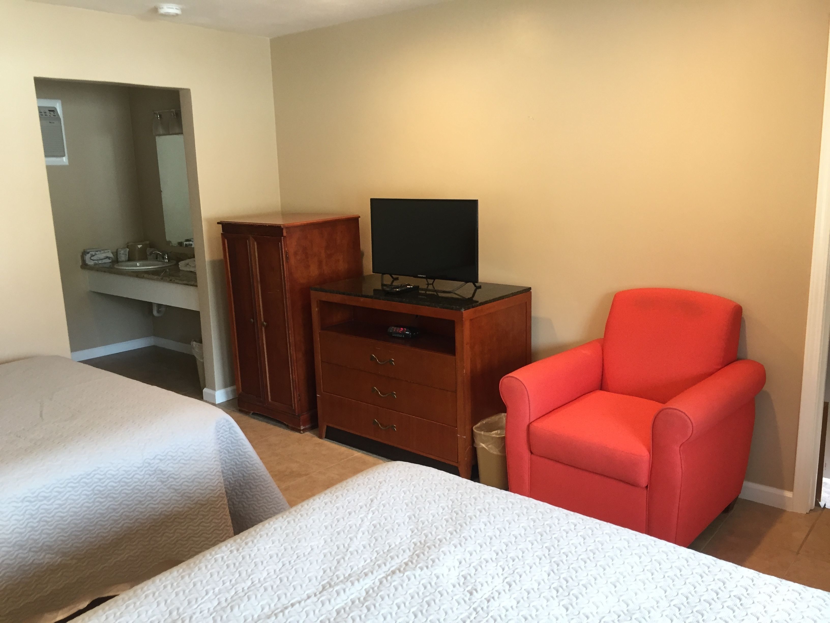 Une chambre d’hôtel avec deux lits, une commode en bois surmontée d’un téléviseur à écran plat, un fauteuil rouge et une salle de bain avec un lavabo et un miroir.