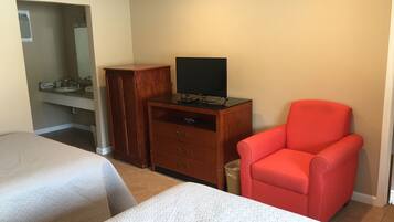 Una habitación de hotel con dos camas, un aparador de madera con un televisor de pantalla plana, una silla roja y un baño con lavamanos y espejo.