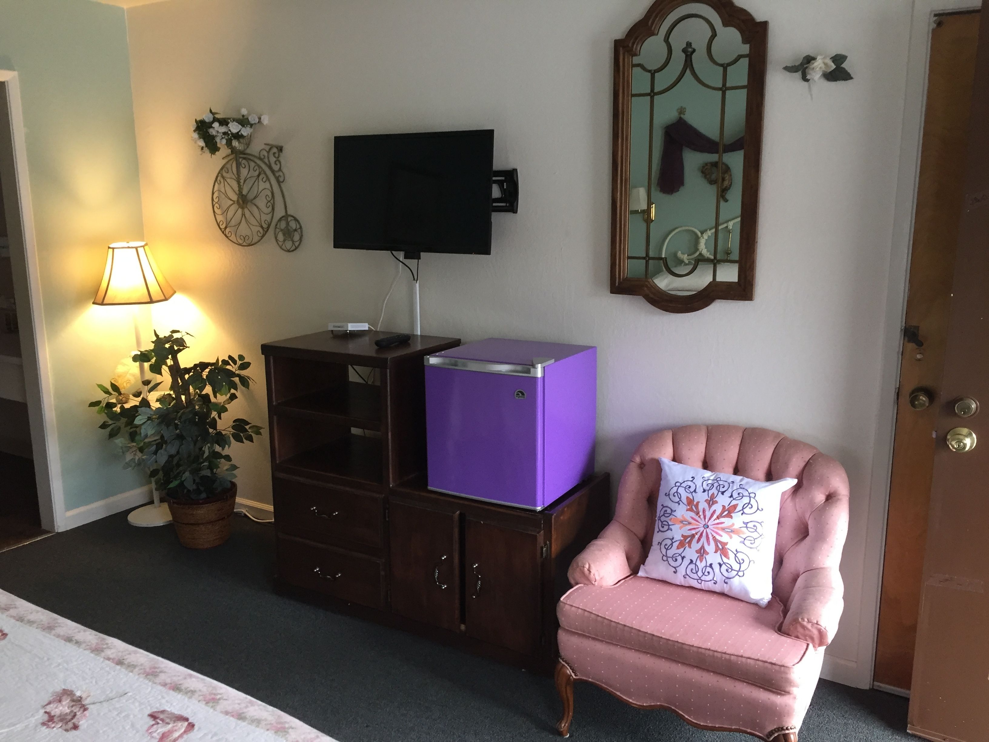 A room with a purple refrigerator, a pink armchair with a patterned pillow, a wall-mounted TV, a potted plant, and a lamp.