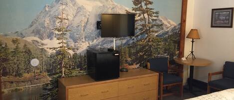A hotel room with a mountain mural, a TV on a wooden dresser, a desk with a lamp, and a bed with a floral bedspread.