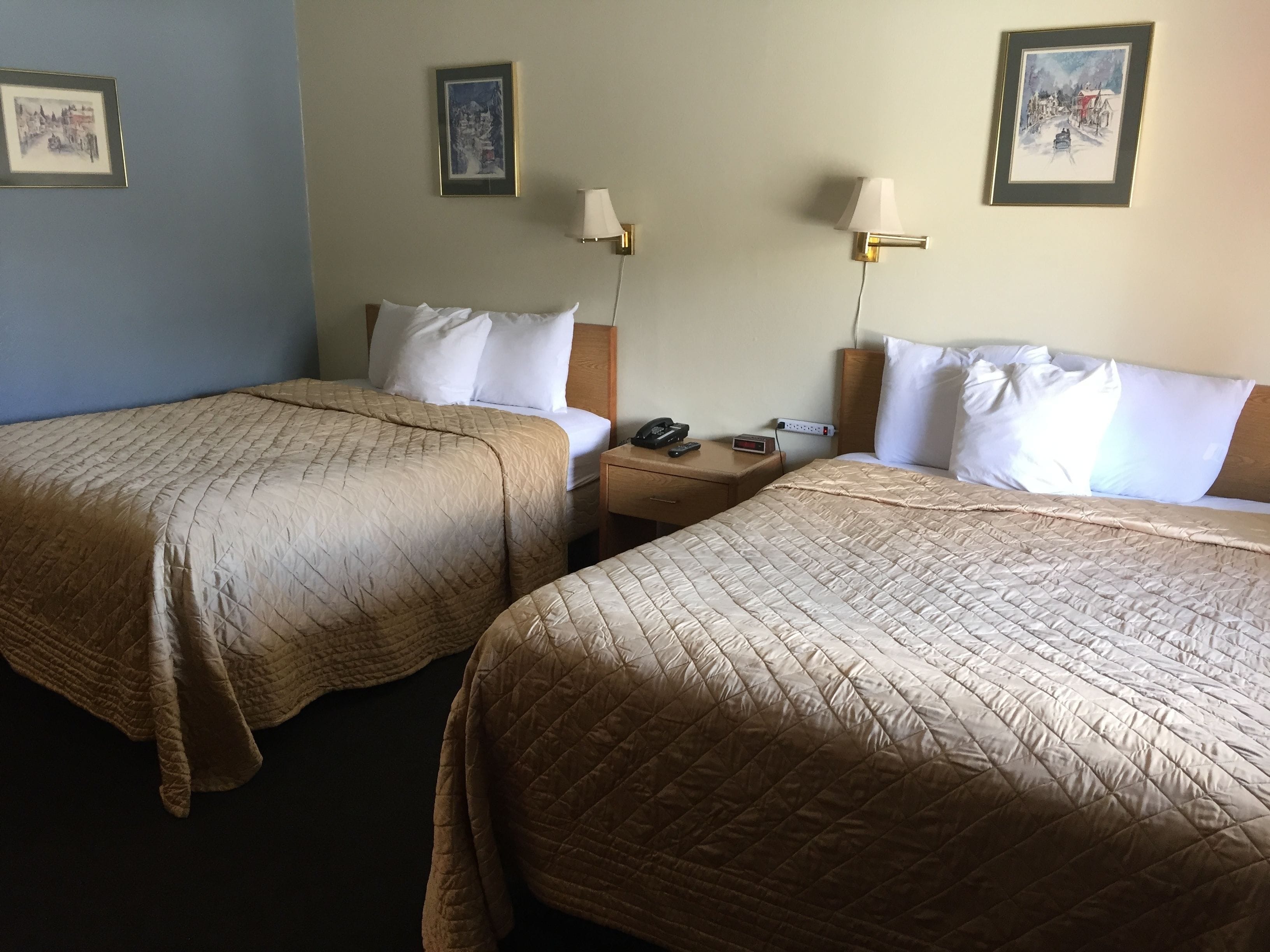 Ein Hotelzimmer mit zwei Betten, jeweils mit einer beigefarbenen gesteppten Bettdecke, weißen Kissen und einem Holzkopfteil. Es gibt zwei Nachttische mit Lampen, einer Uhr und einem Telefon.