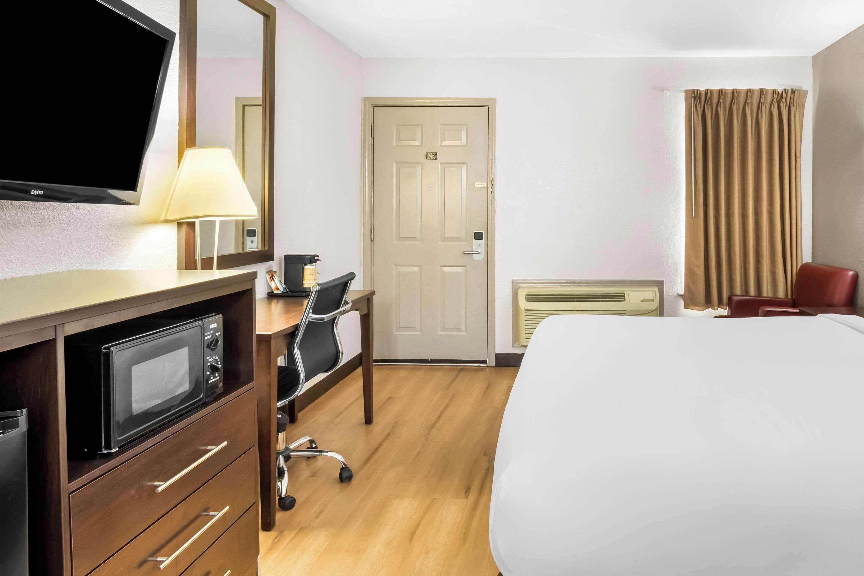 Une chambre d’hôtel avec un lit, un bureau avec un ordinateur, un four à micro-ondes, une lampe, une porte et une fenêtre avec des rideaux.