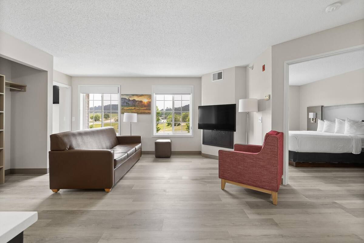 Ein modernes Wohnzimmer mit braunem Sofa, einem roten Sessel, einem Flachbildfernseher und einem Bett mit weißer Bettwäsche.