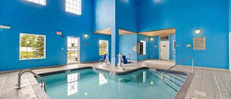 Una piscina interior con paredes azules y techo de madera, que cuenta con ventanas amplias, una silla de salvavidas y una escalera de piscina.