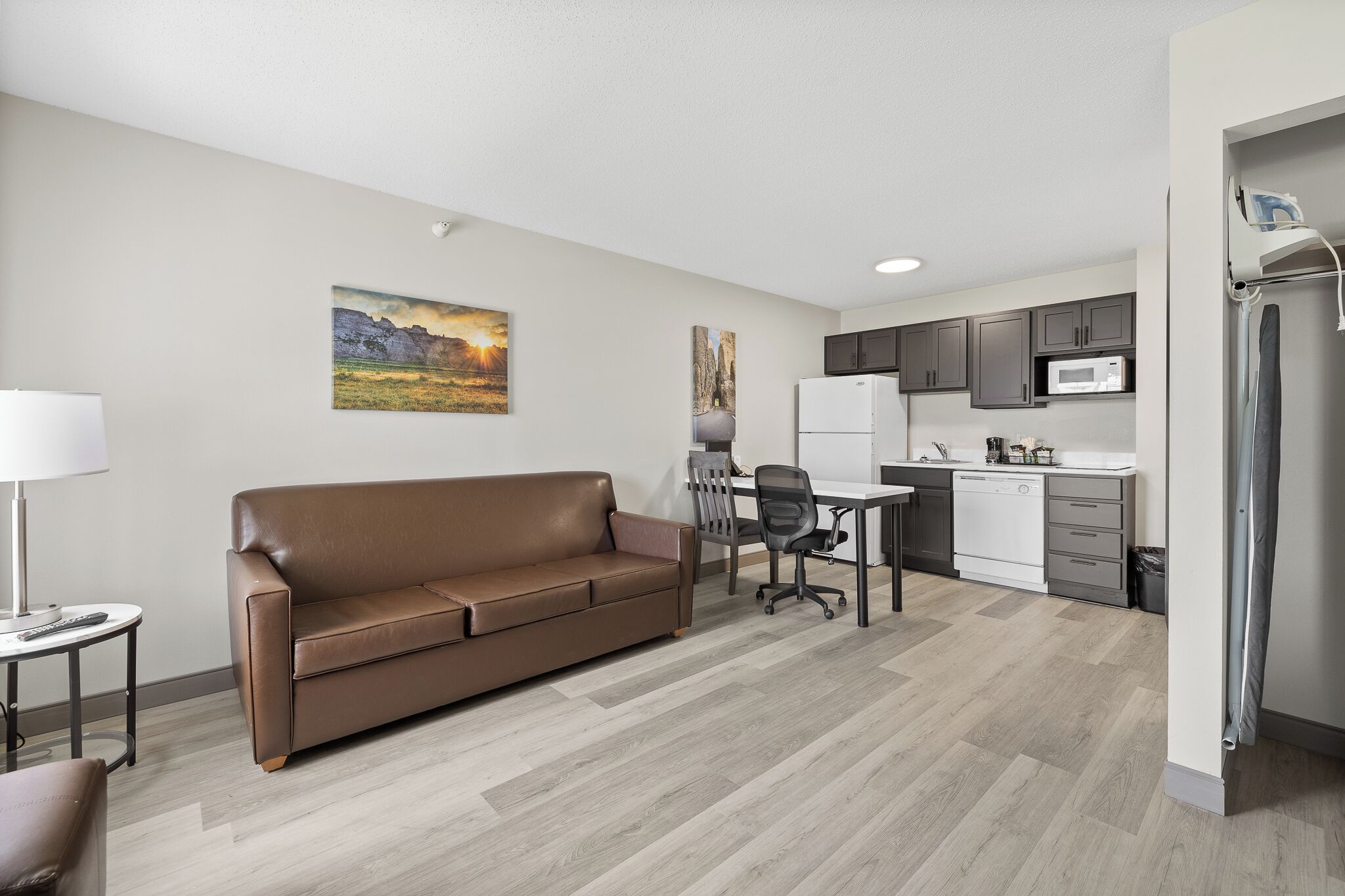A modern apartment with a brown leather sofa, a dining area with a table and chairs, a kitchen with a microwave and refrigerator, and a painting on the wall.