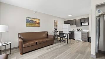 A modern apartment with a brown leather sofa, a dining area with a table and chairs, a kitchen with a microwave and refrigerator, and a painting on the wall.