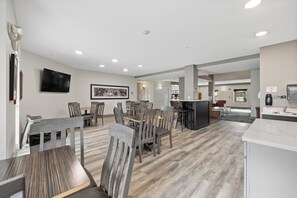 A spacious dining area with wooden tables and chairs, a flat-screen TV mounted on the wall, and a kitchen area with a countertop and sink.