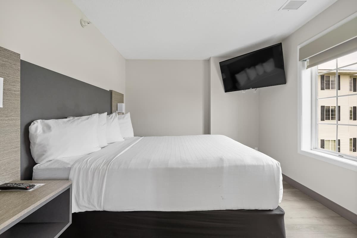 Ein modernes Hotelzimmer mit einem großen Bett, einem an der Wand befestigten Flachbildfernseher, einem Nachttisch und einem Fenster mit Blick auf das Gebäude.