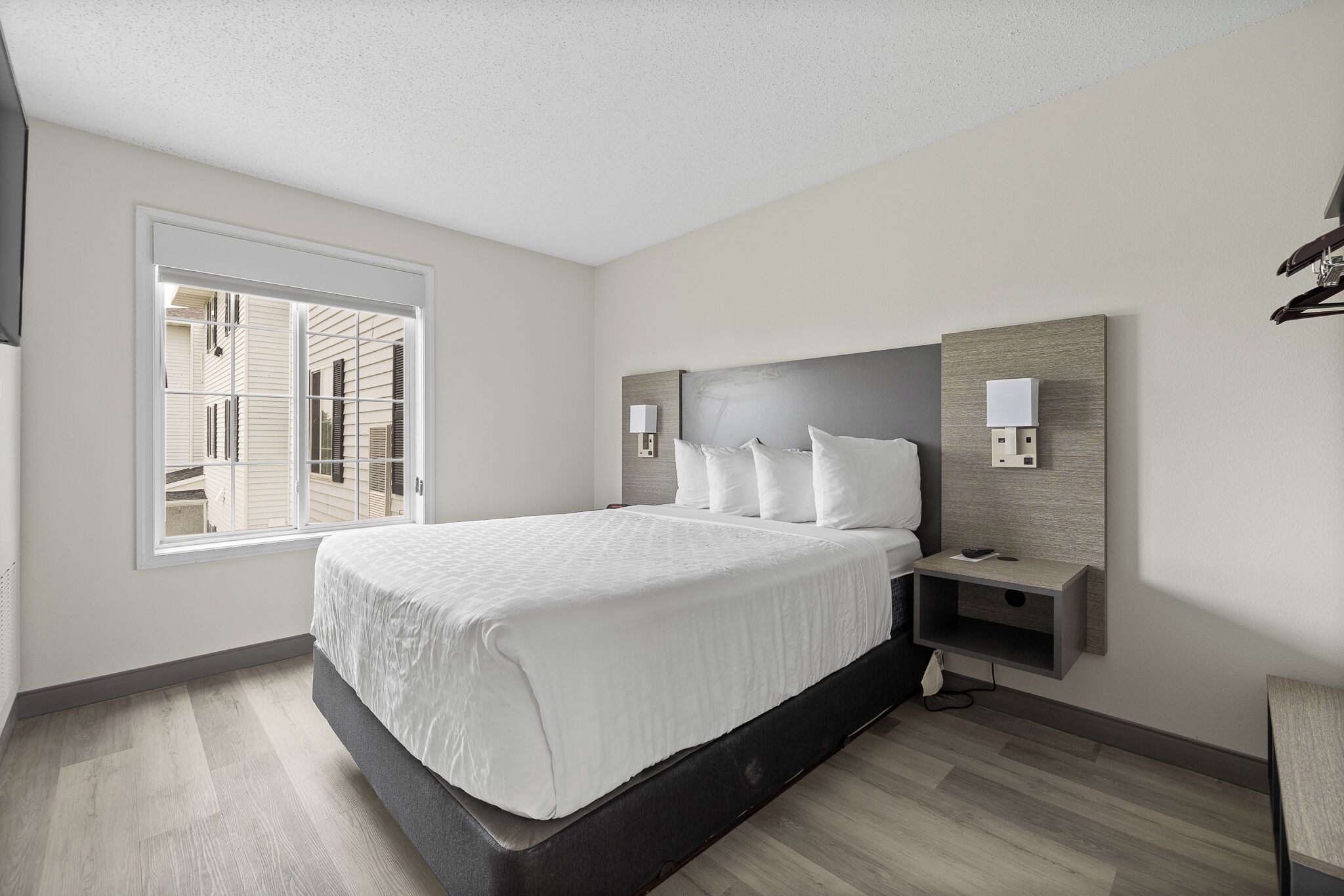 A modern hotel room with a large bed, bedside tables, a wall-mounted TV, and a window with a view of neighboring buildings.