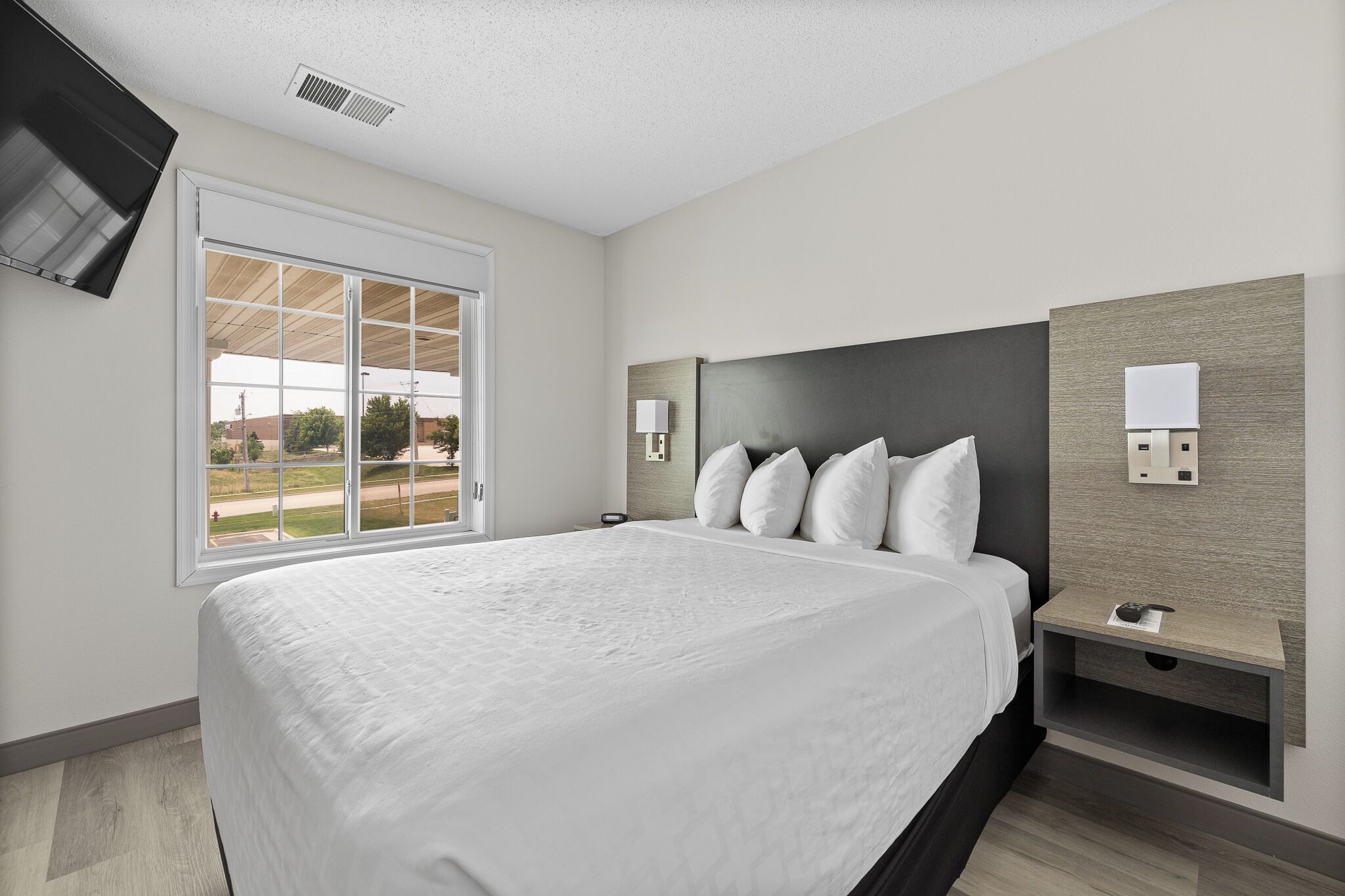 A modern hotel room with a large bed, a sliding glass door to the outside, a flat-screen TV mounted on the wall, and a bedside table with a lamp.