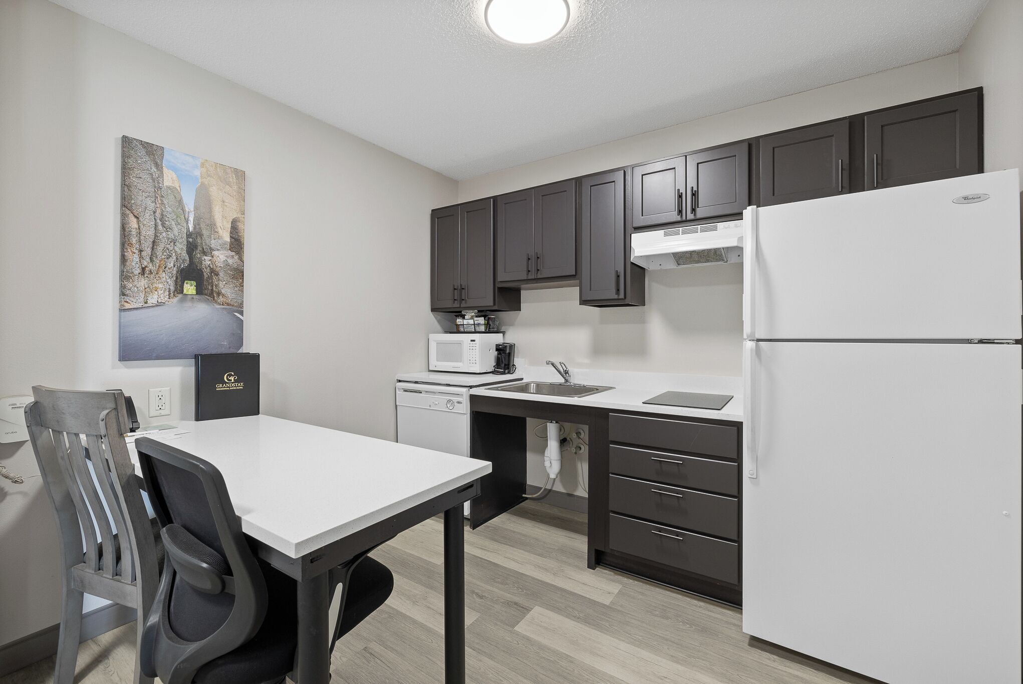 A modern kitchen with dark cabinets, a white refrigerator, a microwave, and a dining area with a white table and chairs.