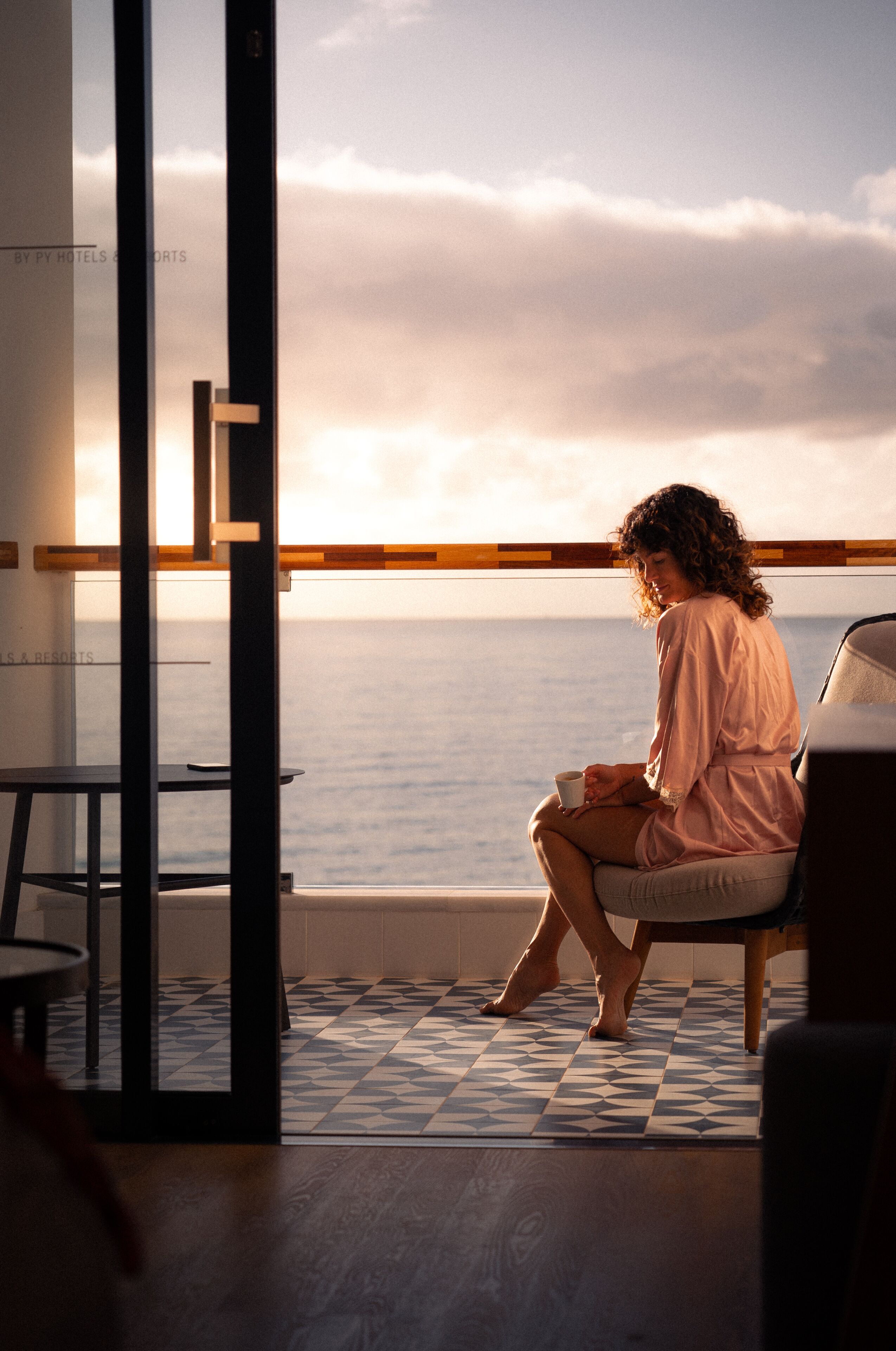 Una persona sentada junto a una ventana con vistas al mar, donde se aprecia el océano y un piso de baldosas.