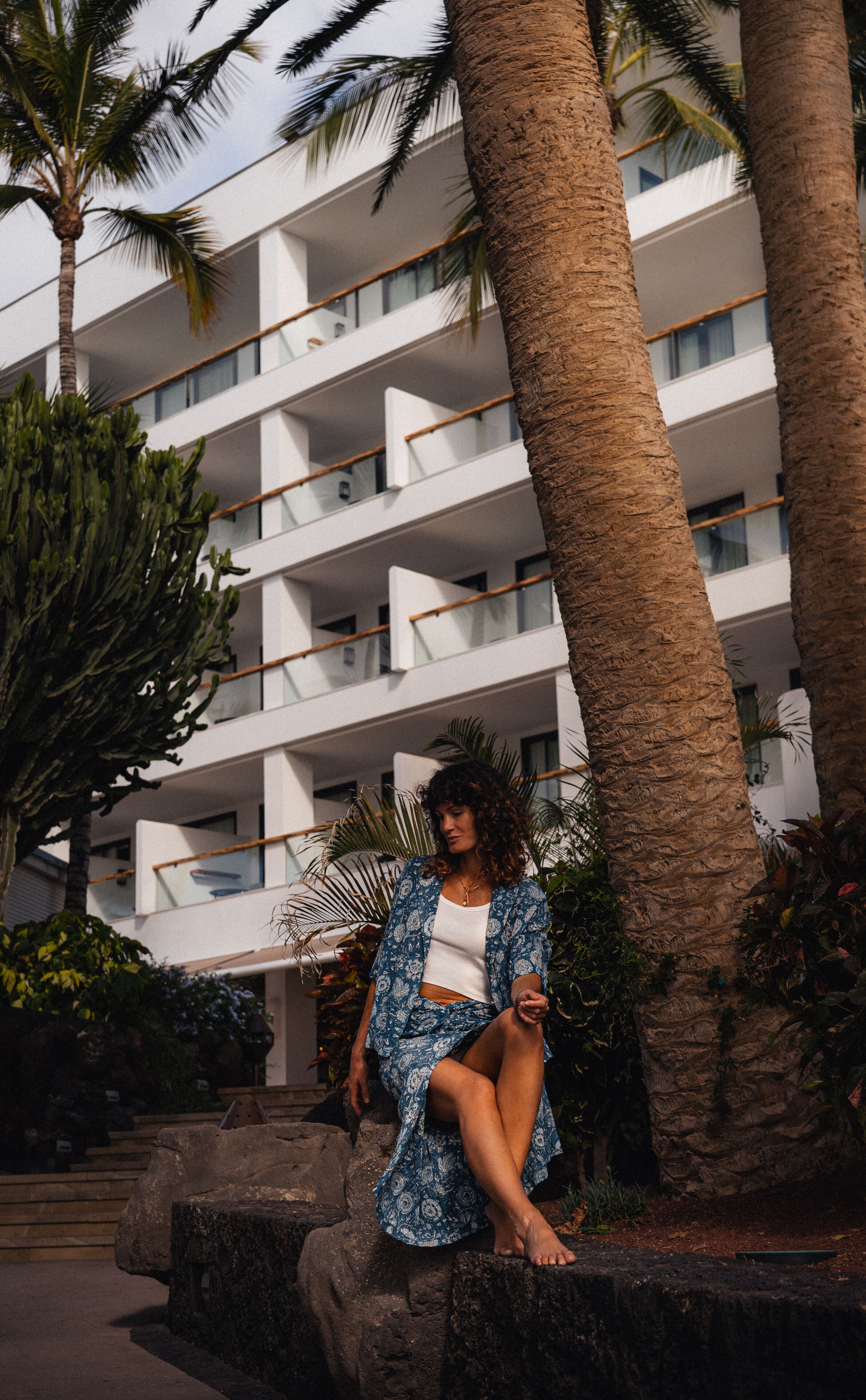 A person sitting on a stone ledge in front of a multi-story building with balconies, surrounded by palm trees and other vegetation.