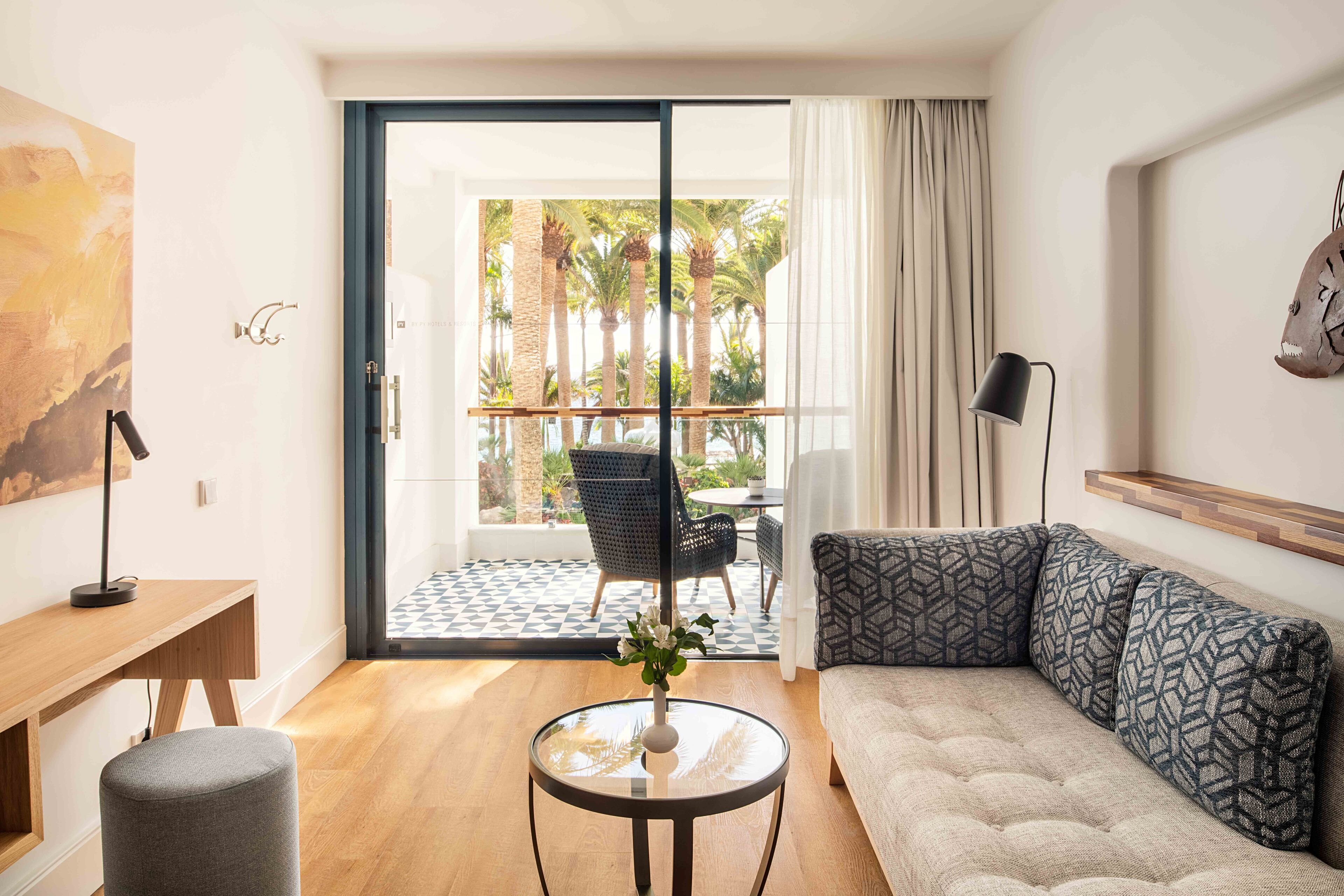 A modern living room with a sofa, a small round table with a vase, a desk, and a chair. There is a large window with a view of palm trees and a balcony.