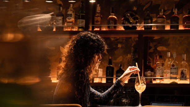 Une personne assise au bar, un verre à la main, entourée de bouteilles, dans un intérieur chaleureux et faiblement éclairé.