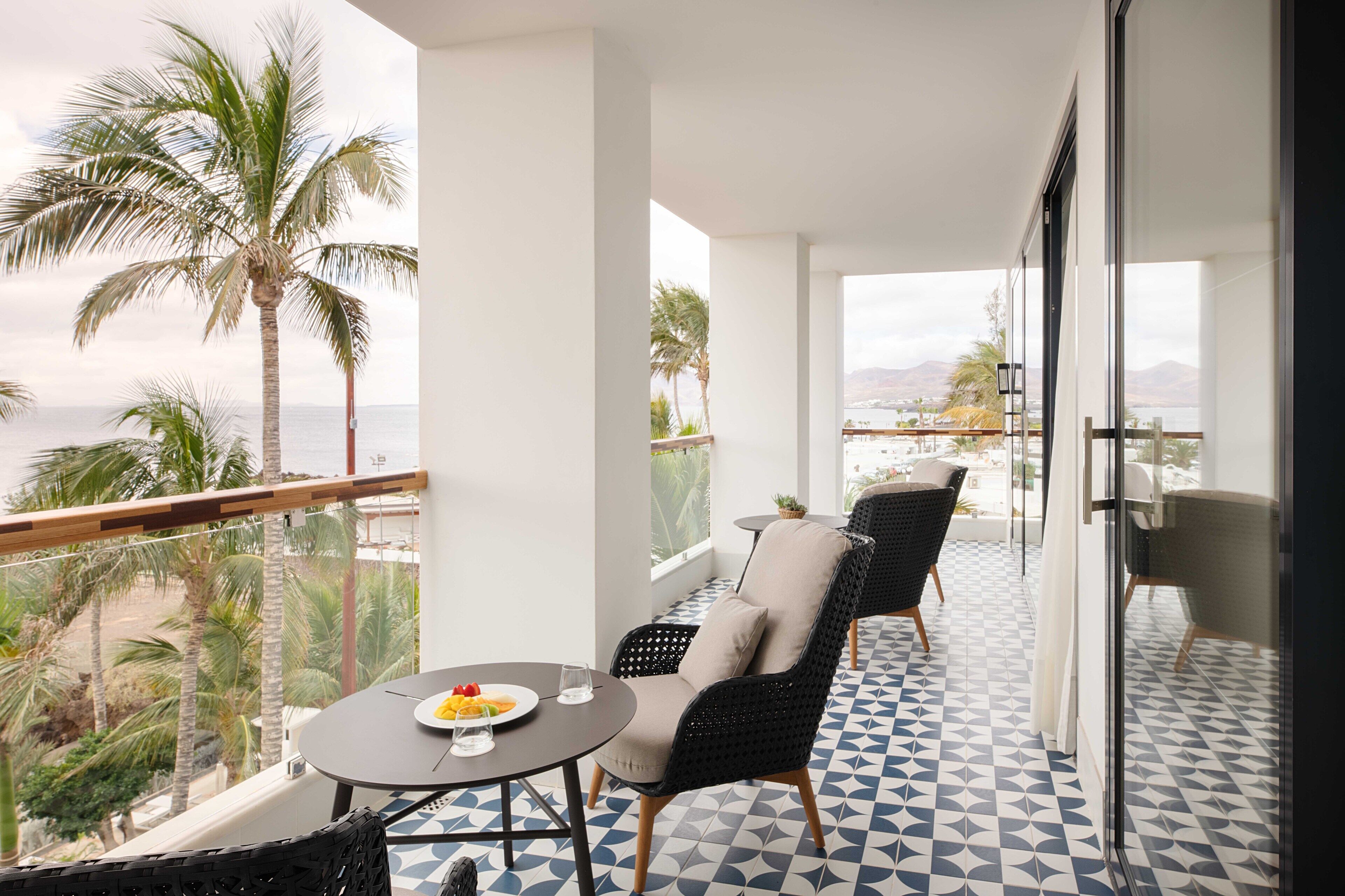 A balcony with a table, chairs, and a view of palm trees and the sea.