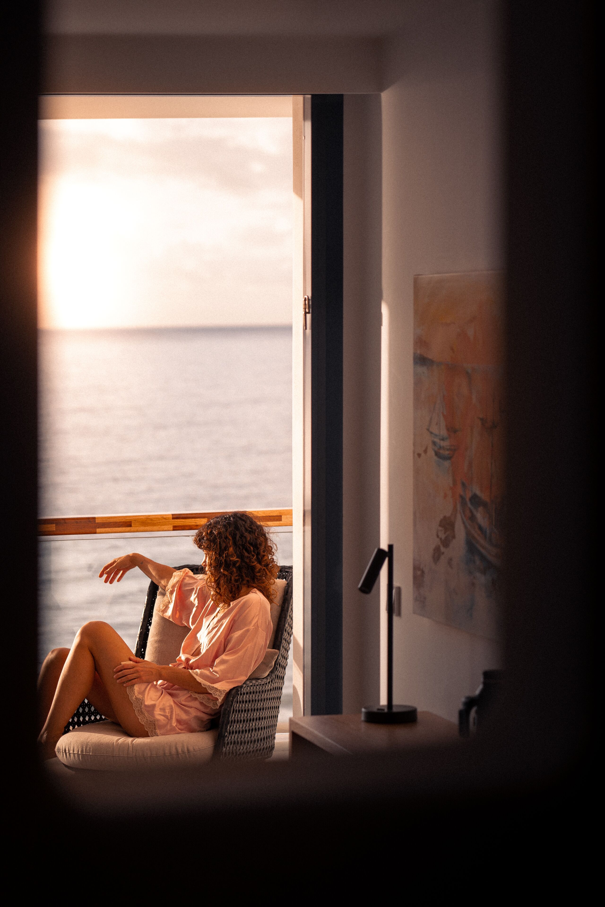 Una persona sentada junto a una ventana con vista al mar, con una silla y una lámpara sobre una mesa.