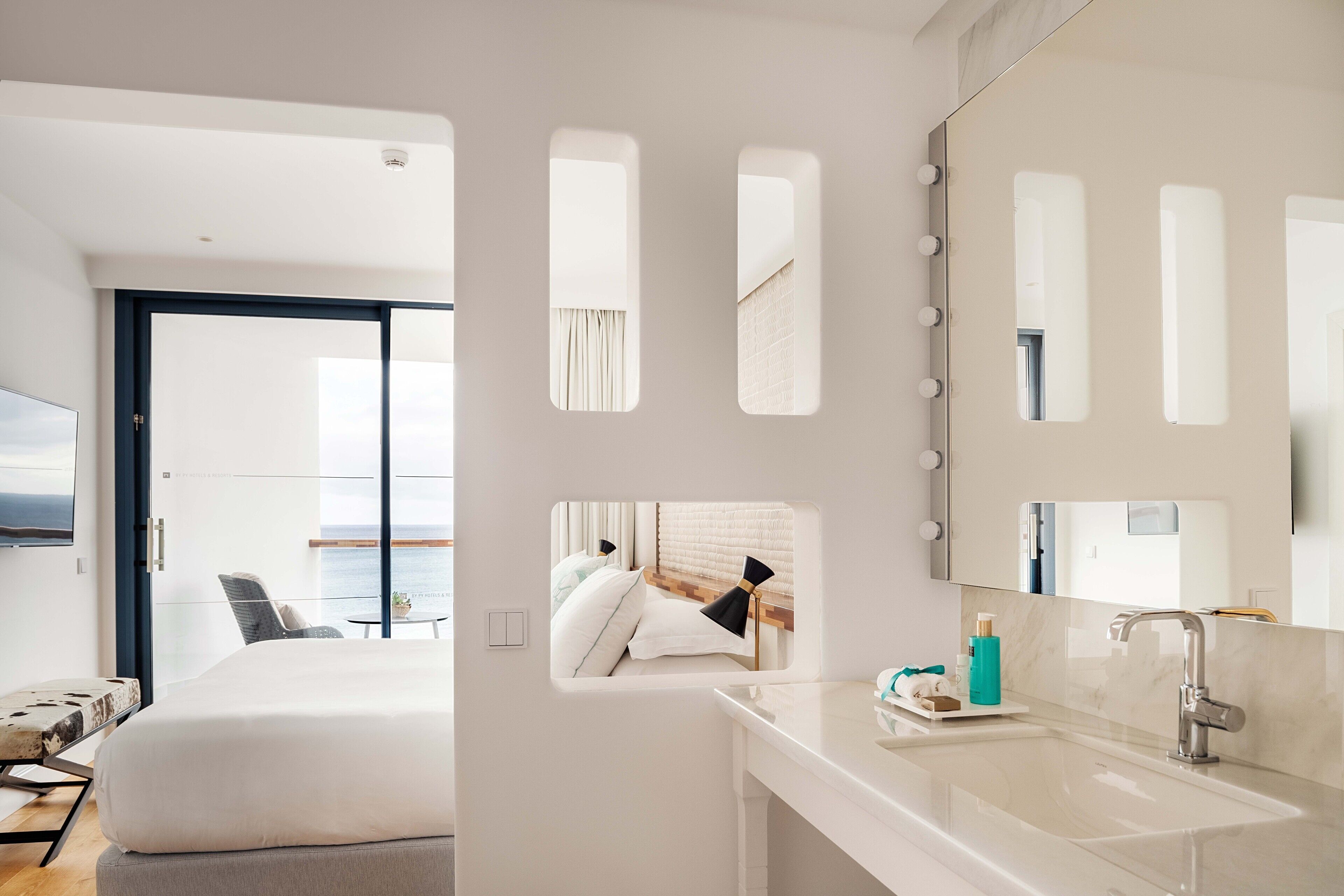 A modern hotel room with a large bed, a bathroom with a mirror and sink, and a balcony with a view.