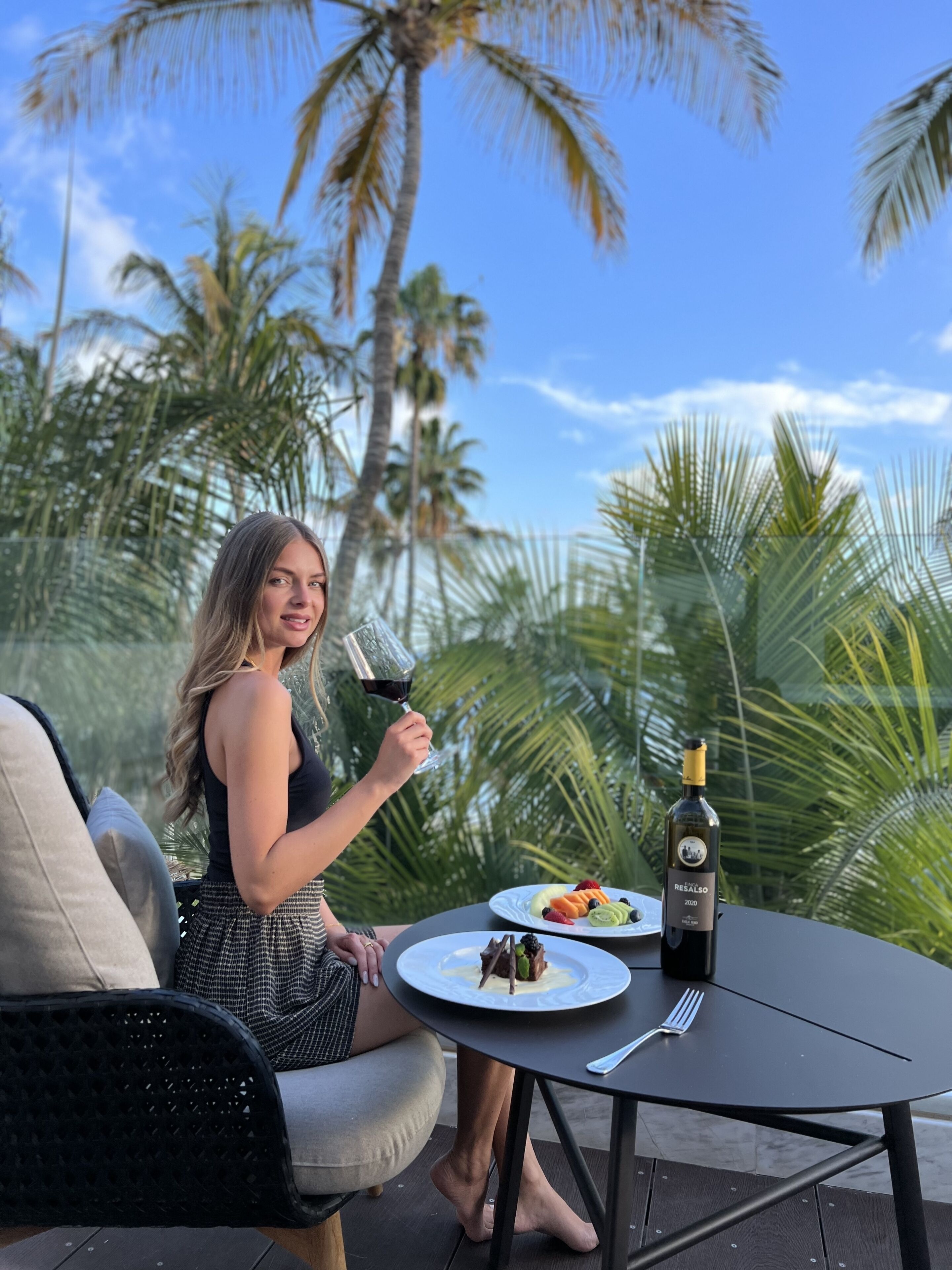 Uma mulher jantando ao ar livre, com uma mesa posta para dois, uma garrafa de vinho e um prato de comida.
