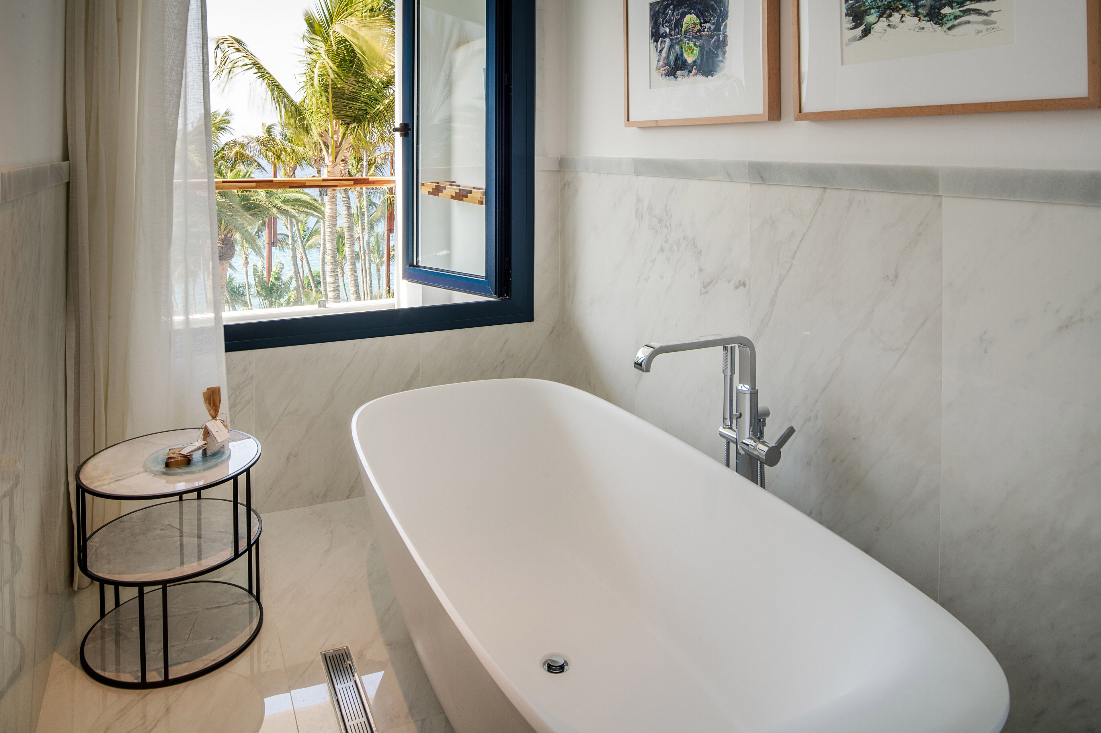 Un baño moderno con una bañera independiente, una pequeña mesa redonda con un florero y una ventana con vista a palmeras.