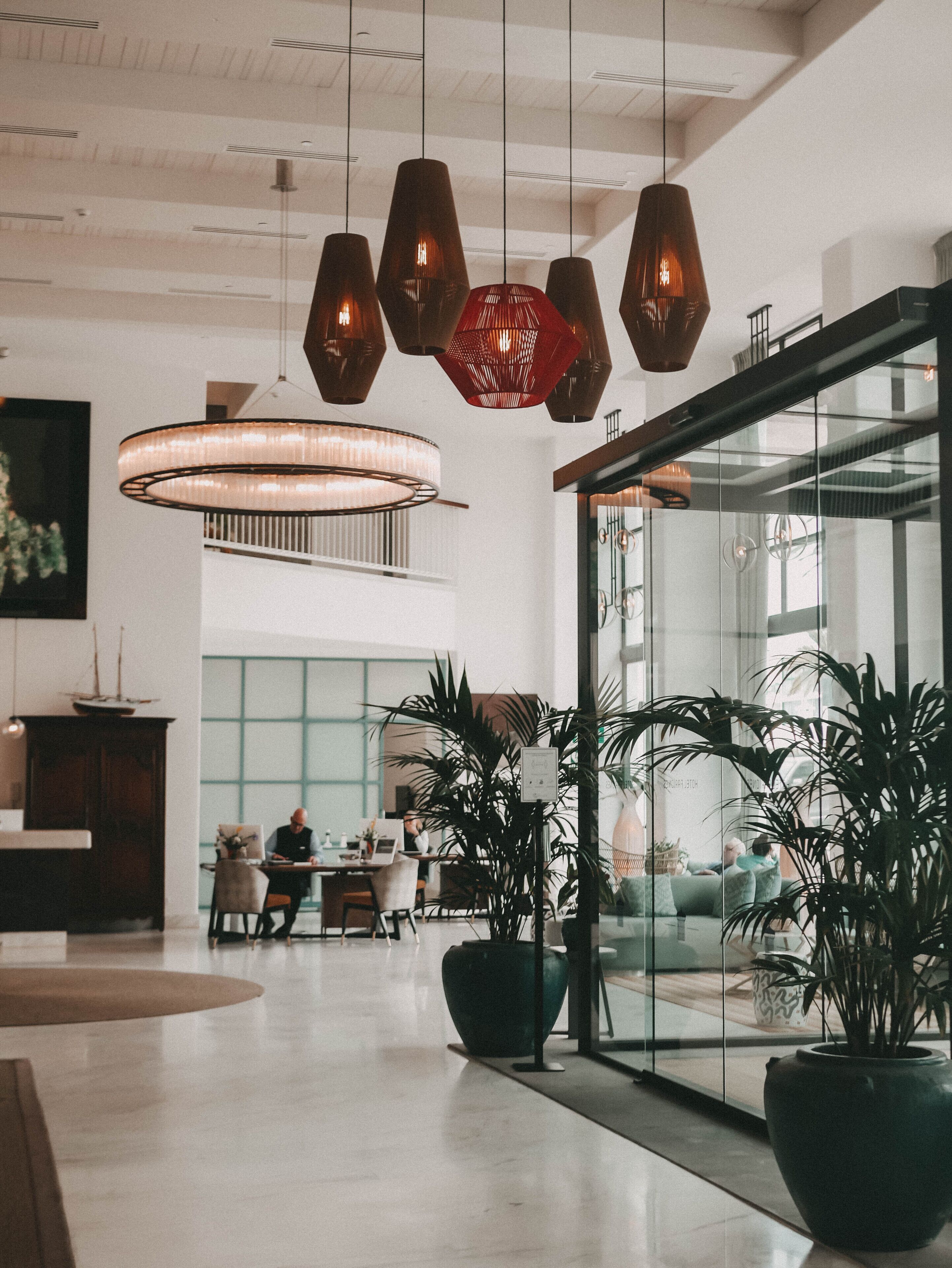 A spacious lobby with modern pendant lighting, large potted plants, and a seating area with a glass wall offering views of the interior.