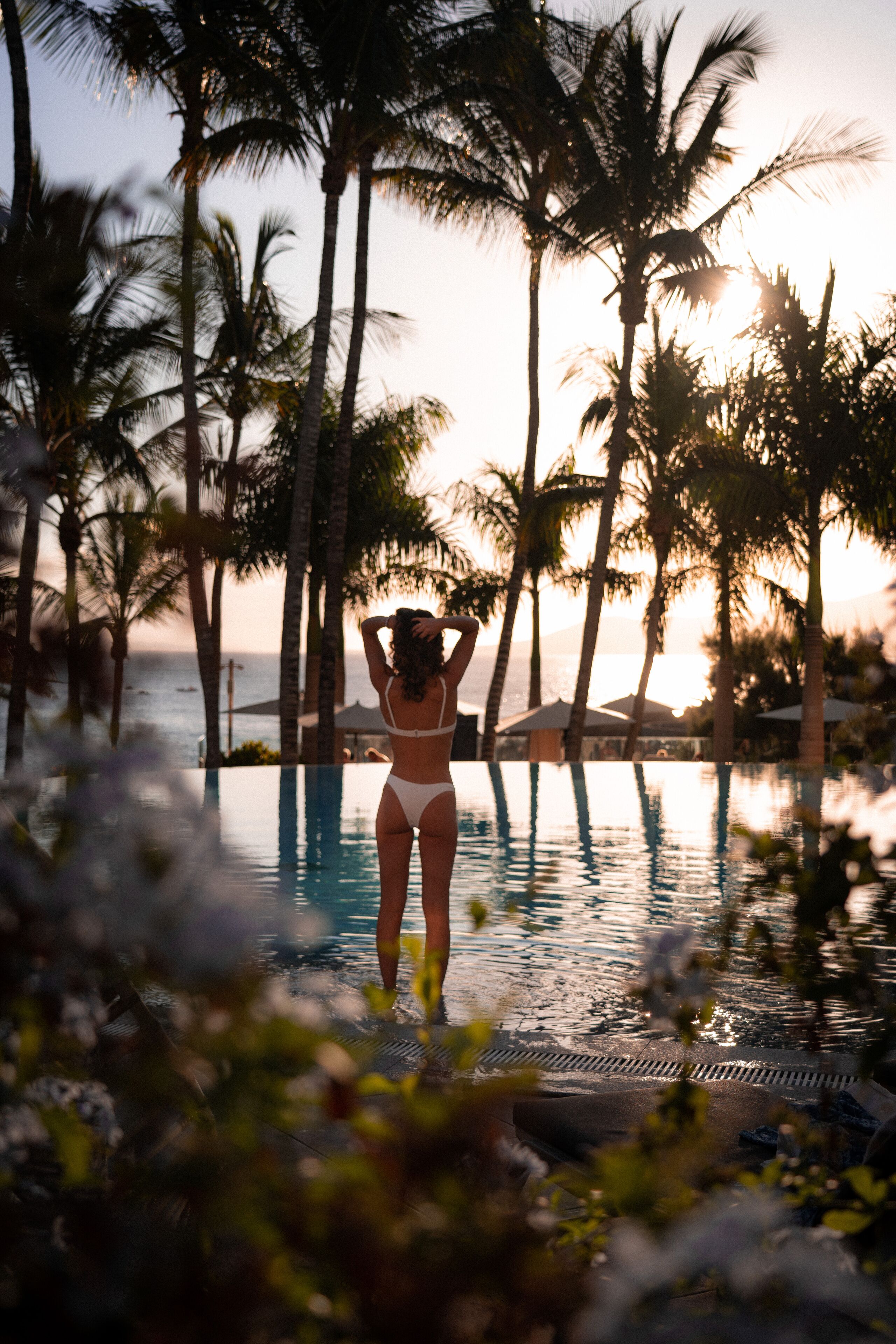 Une personne vêtue d’un bikini blanc, debout dans une piscine entourée de palmiers, avec l’océan en toile de fond.