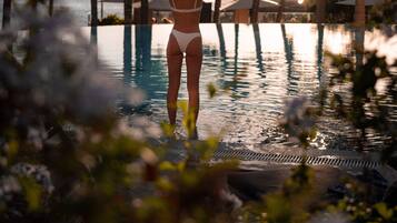 Une personne vêtue d’un bikini blanc, debout dans une piscine entourée de palmiers, avec l’océan en toile de fond.