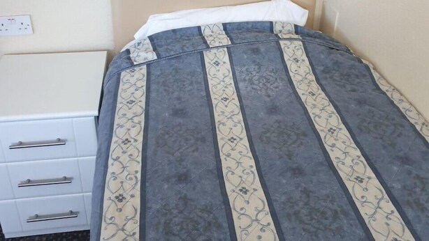 A small, single-bed room with a plain headboard, a white bedside table with drawers, and a patterned bedspread. - Elmhurst Hotel (Reading)